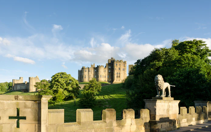 Alnwick Castle viewed from Lion Bridge with stone lion statue in foreground.