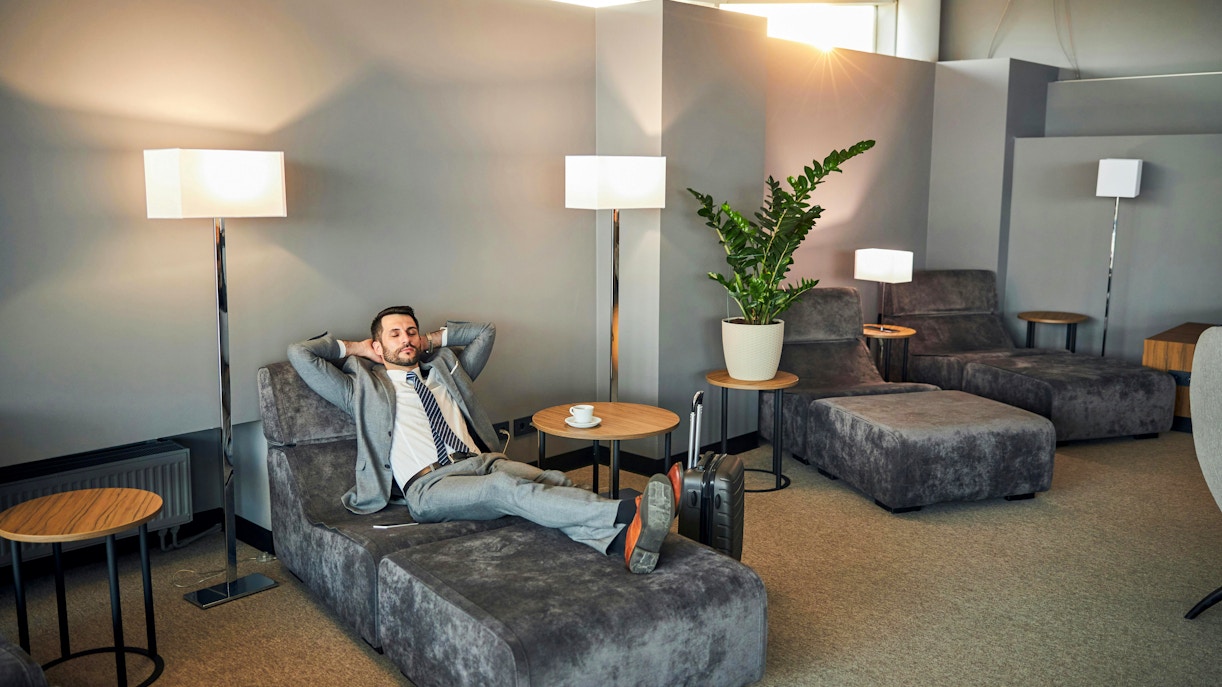 Man relaxing on a lounge chair in an airport rest area with suitcase nearby.