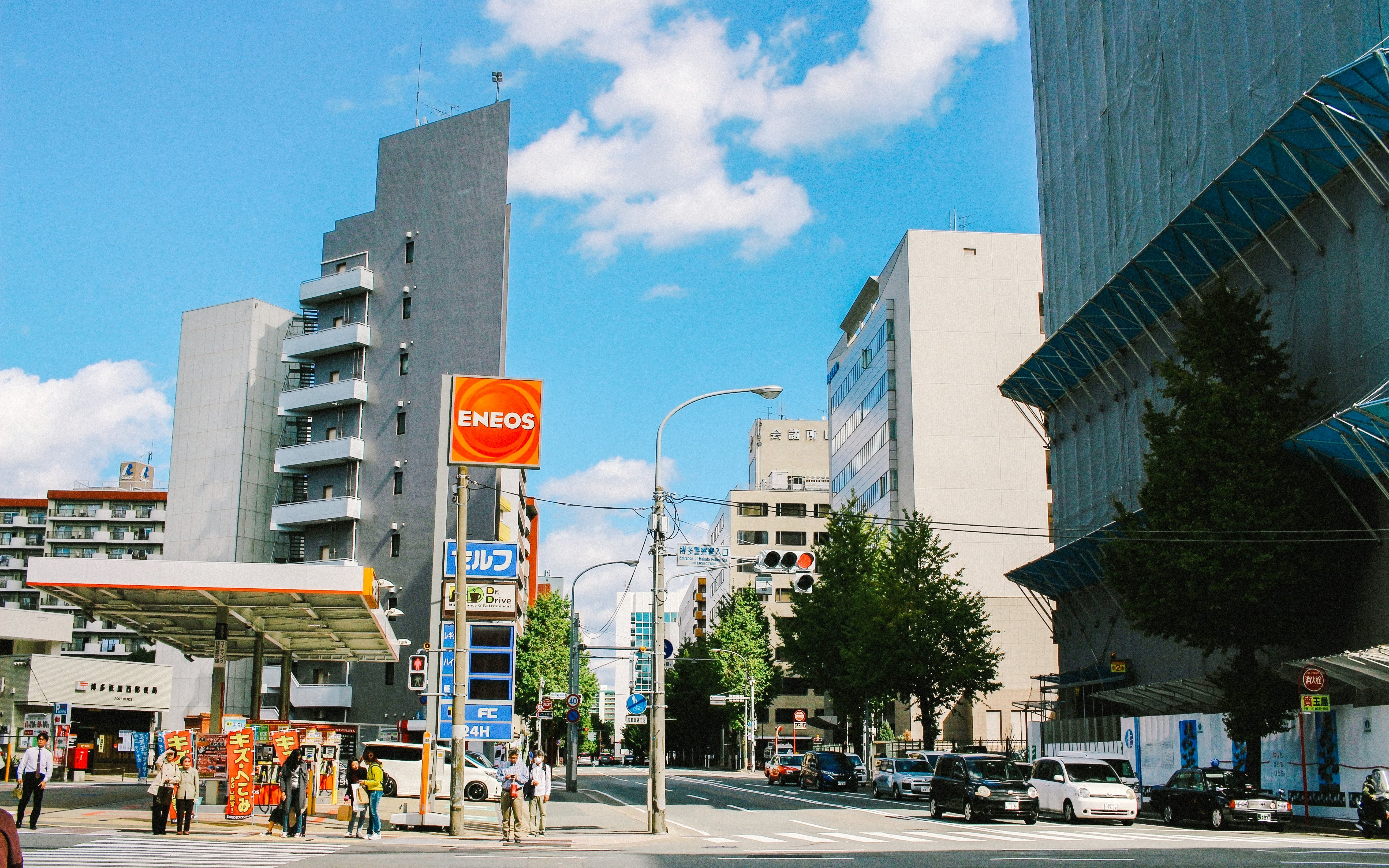 ENEOS gas station on a city street with surrounding buildings and pedestrians.