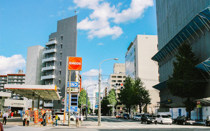 ENEOS gas station on a city street with surrounding buildings and pedestrians.