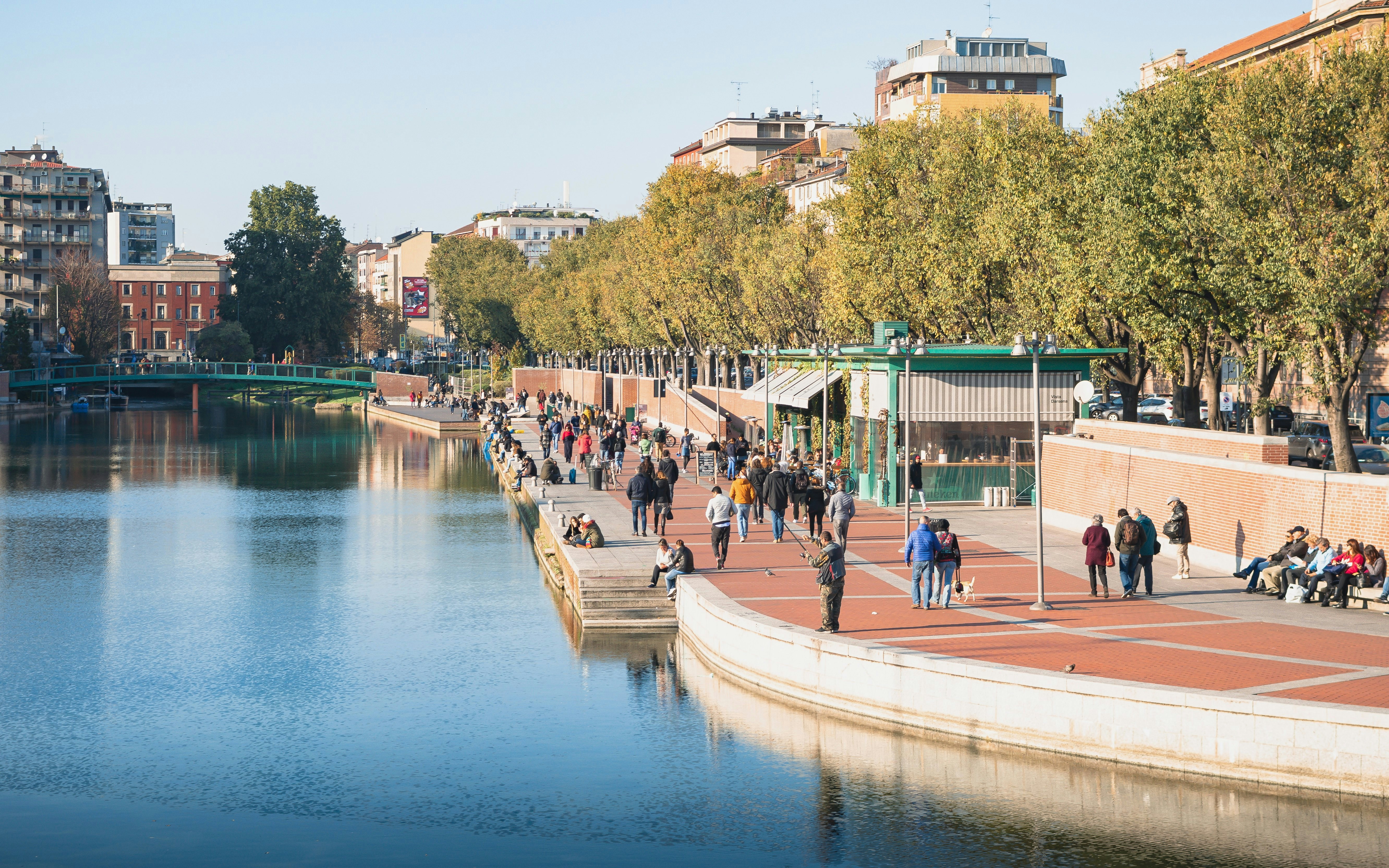 People walking along Darsena dock in Milan with trees and buildings in the background.