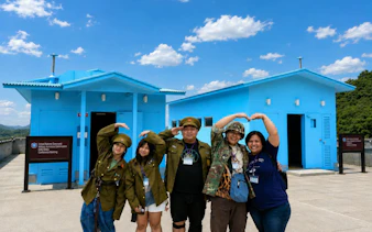 Visitors in military attire pose at the DMZ's JSA Photo Zone, near blue conference buildings.