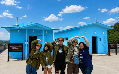 Visitors in military attire pose at the DMZ's JSA Photo Zone, near blue conference buildings.