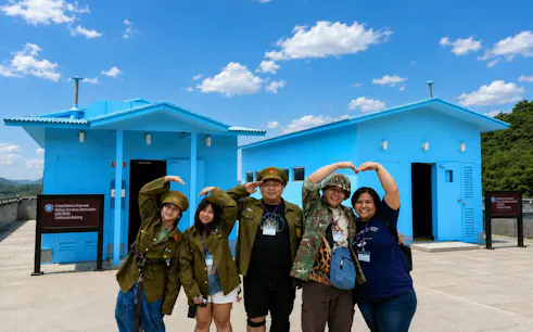 Visitors in military attire pose at the DMZ's JSA Photo Zone, near blue conference buildings.