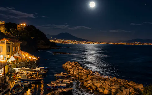 Naples coastline at night with boats, illuminated buildings, and Mount Vesuvius in the background.