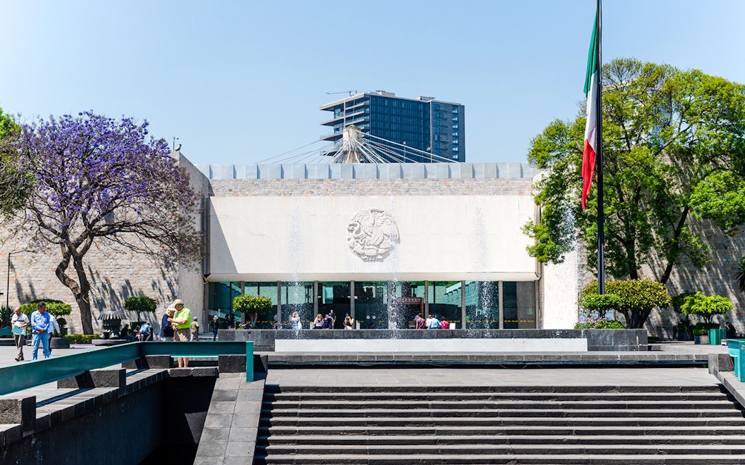 Main entrance of the Anthropology Museum in Mexico City with visitors and Mexican flag.