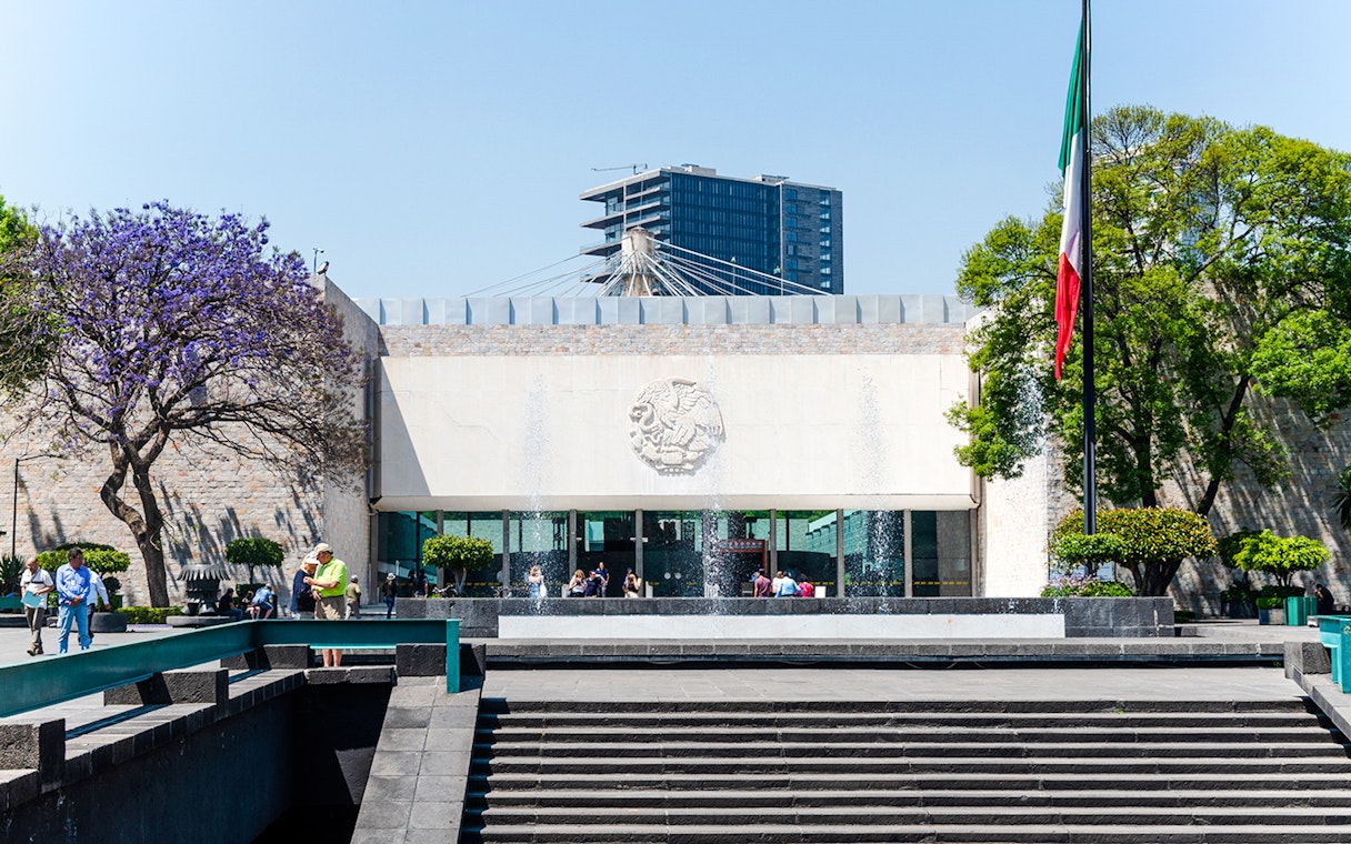 Main entrance of the Anthropology Museum in Mexico City with visitors and Mexican flag.