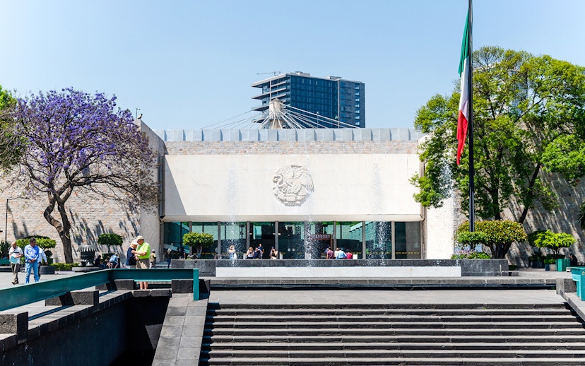 Main entrance of the Anthropology Museum in Mexico City with visitors and Mexican flag.