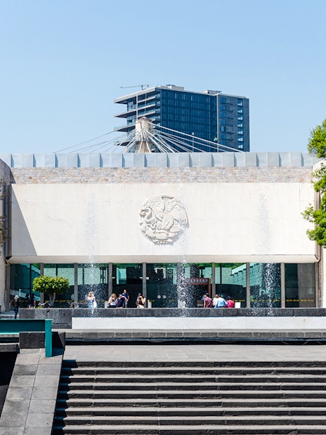Main entrance of the Anthropology Museum in Mexico City with visitors and Mexican flag.