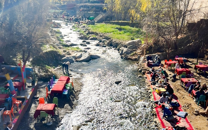 Outdoor dining by a river in Ourika Valley, Morocco, with colorful tables and people enjoying meals.