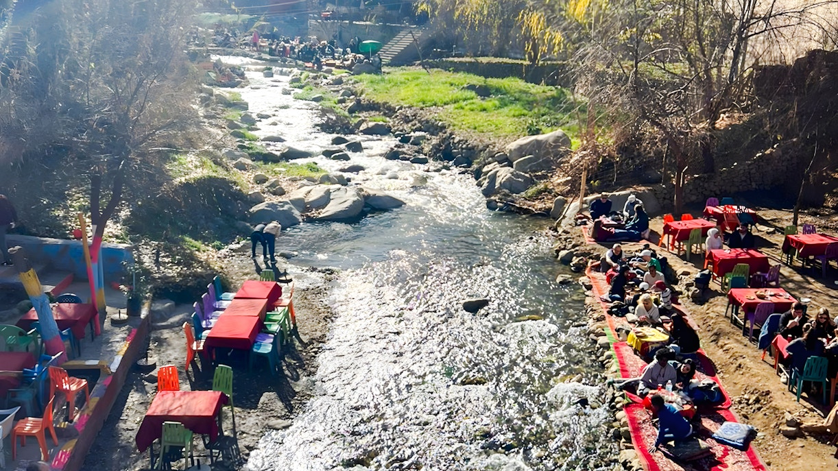 Outdoor dining by a river in Ourika Valley, Morocco, with colorful tables and people enjoying meals.