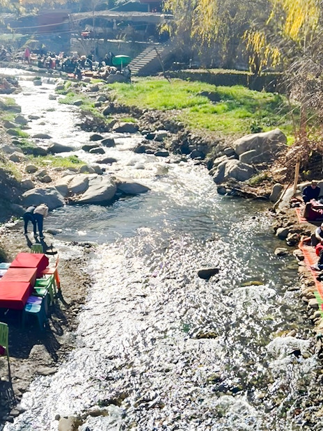 Outdoor dining by a river in Ourika Valley, Morocco, with colorful tables and people enjoying meals.