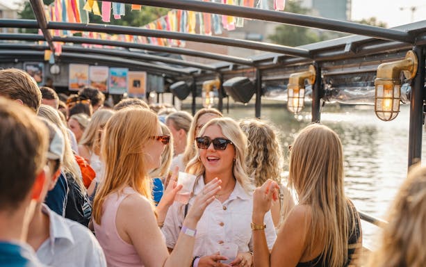 Guests enjoying a lively terrace boat party on a river with colorful decorations.