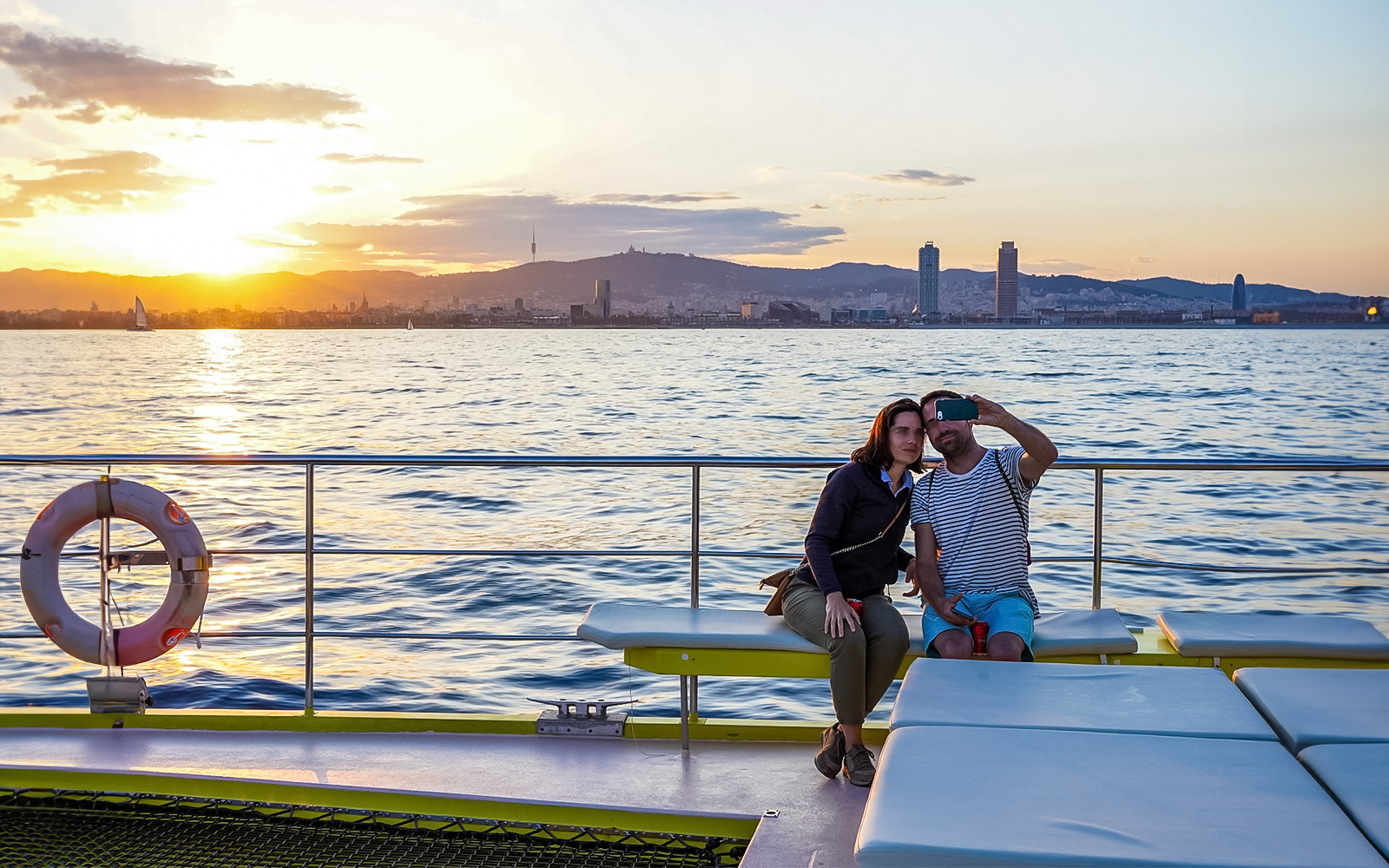 Couple taking selfie on catamaran cruise with Barcelona skyline at sunset.