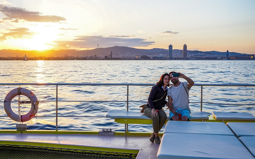 Couple taking selfie on catamaran cruise with Barcelona skyline at sunset.