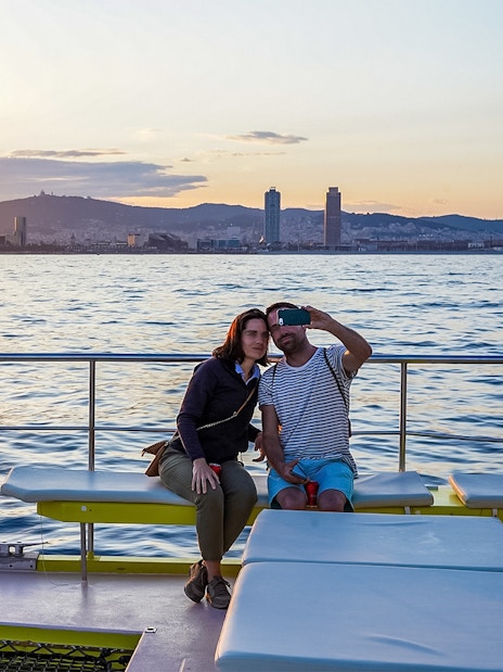 Couple taking selfie on catamaran cruise with Barcelona skyline at sunset.