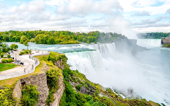 Niagara Falls view from American side with morning mist and tourists on observation path.