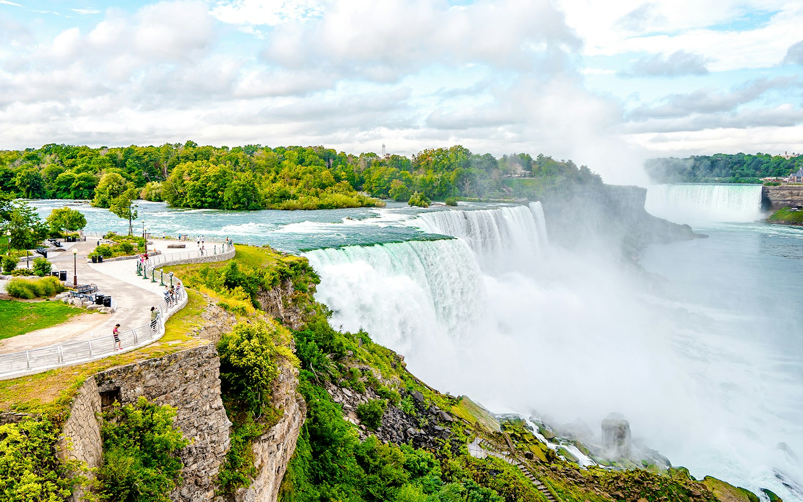 Niagara Falls view from American side with morning mist and tourists on observation path.