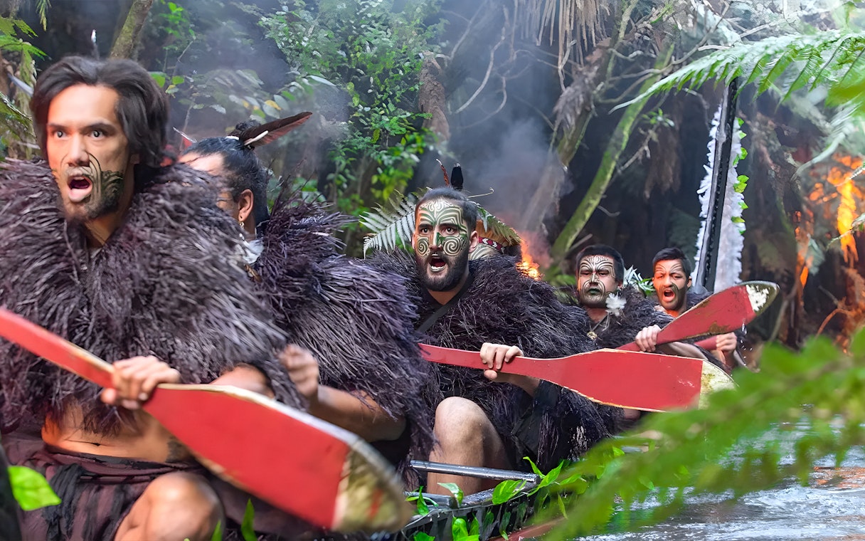 Warriors in traditional attire paddling a canoe on Wai o Whiro stream at Mitai Maori Village.