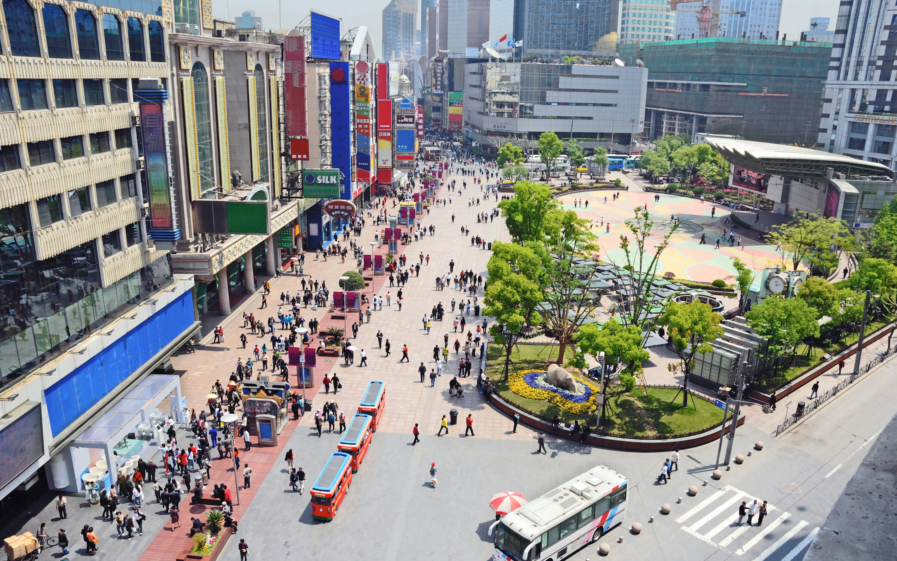 Pedestrians walking along Nanjing Road in Shanghai, China, with colorful buildings and a park area.