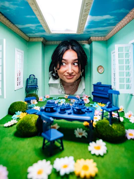 Person peering into a miniature room with blue furniture and floral decor at Studio of Wonders.