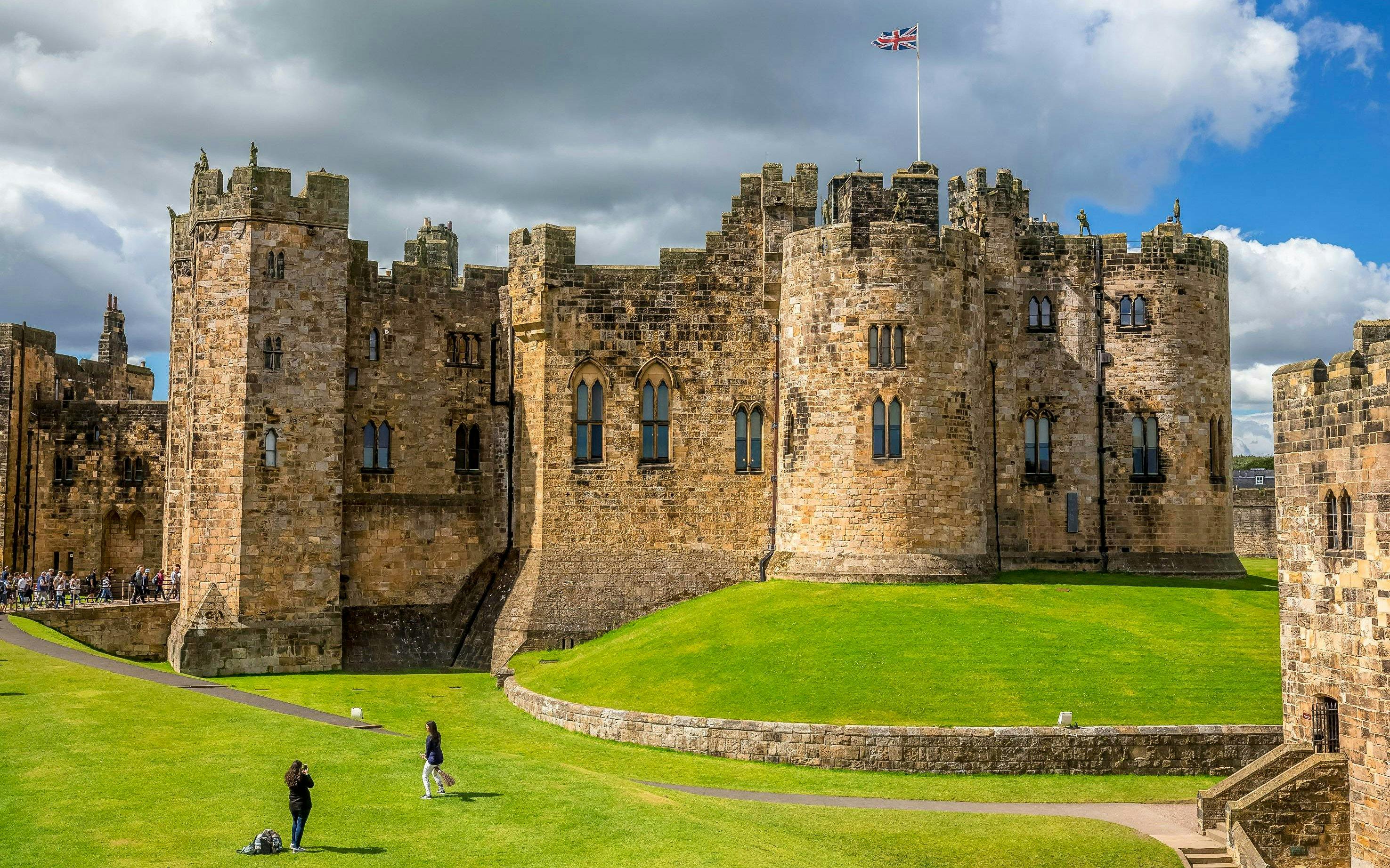 Alnwick Castle exterior with stone towers and green lawns under a cloudy sky.