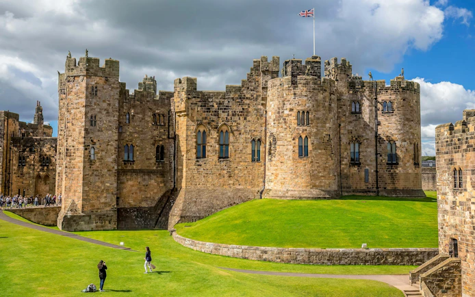 Alnwick Castle exterior with stone towers and green lawns under a cloudy sky.