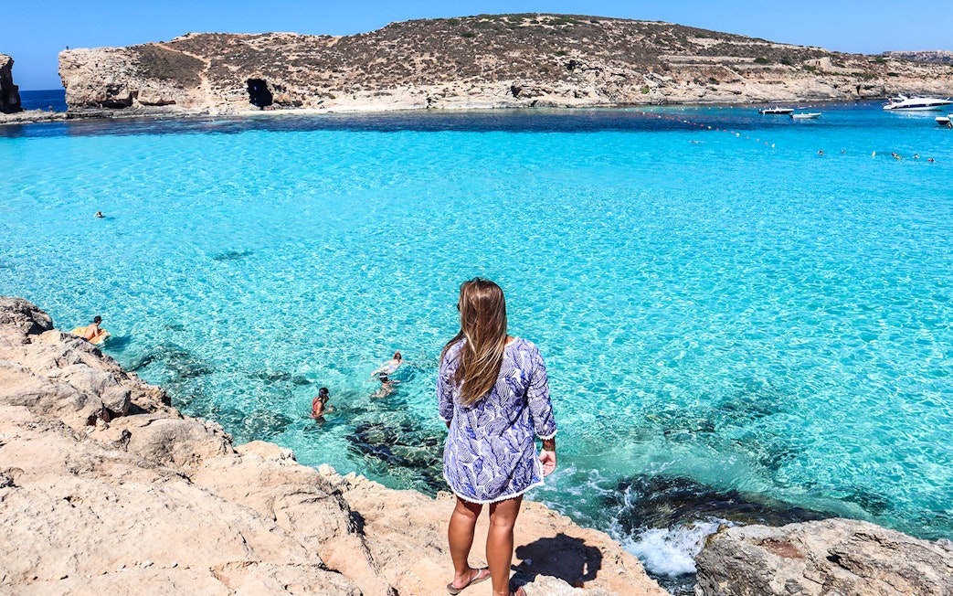 Woman overlooking clear blue waters at Comino, Malta, with swimmers and rocky coastline.