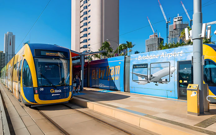 Tram at station with Brisbane Airport advertisement, city buildings in background.