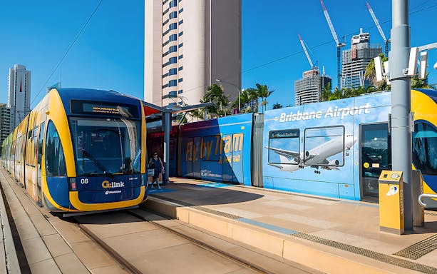 Tram at station with Brisbane Airport advertisement, city buildings in background.