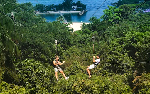 Two people ziplining over lush greenery at Mega Adventure Park, Singapore, with a beach view.