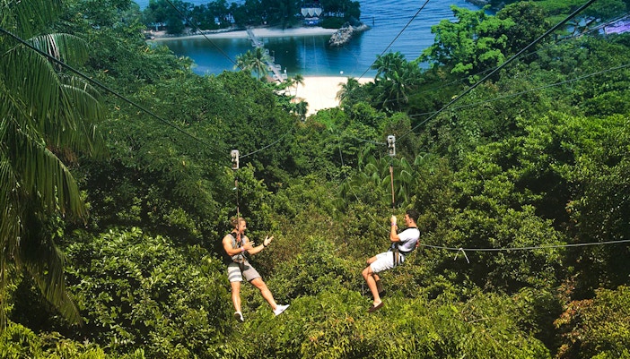 Two people ziplining over lush greenery at Mega Adventure Park, Singapore, with a beach view.