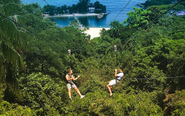Two people ziplining over lush greenery at Mega Adventure Park, Singapore, with a beach view.