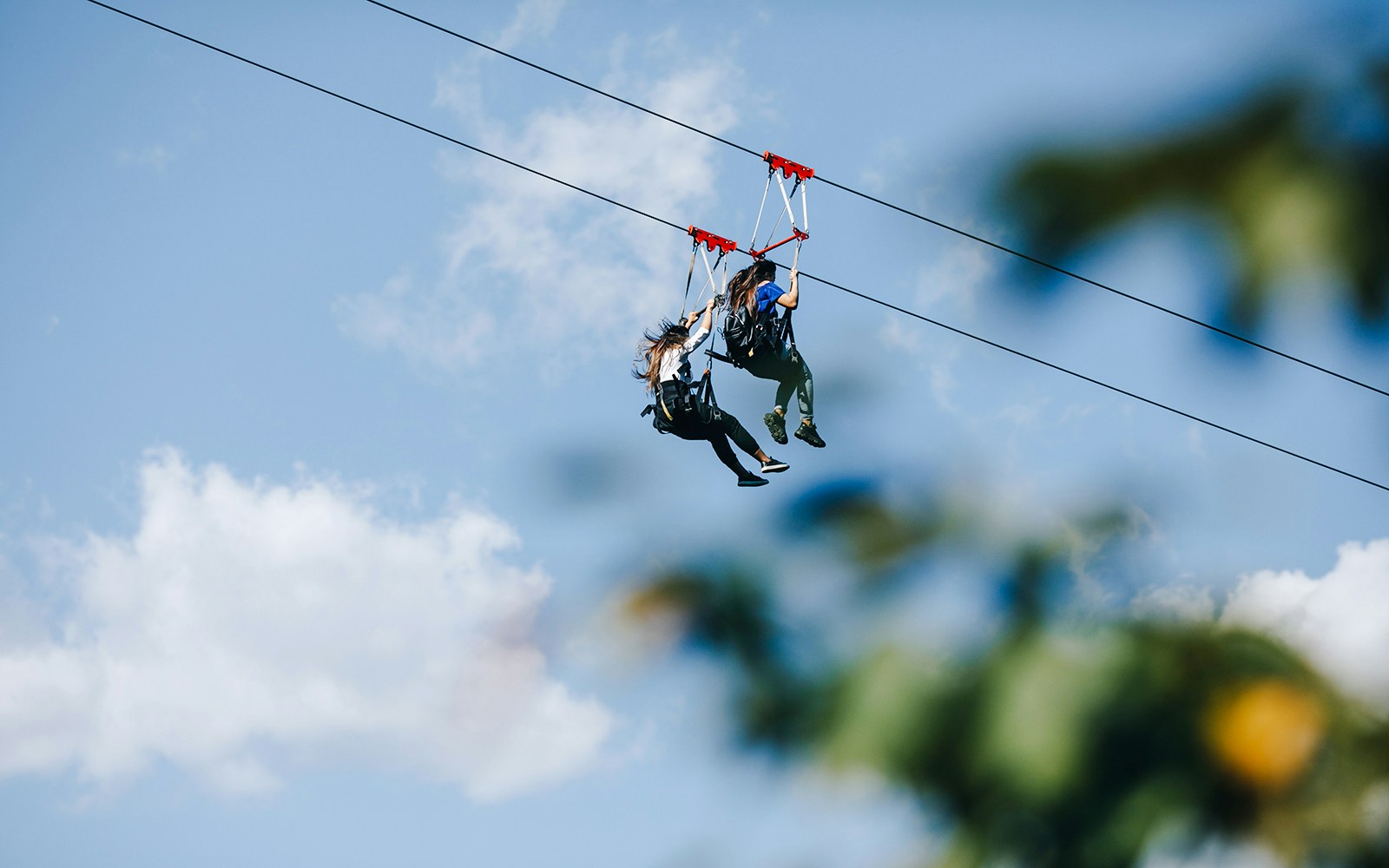 Two people ziplining against a blue sky with clouds.