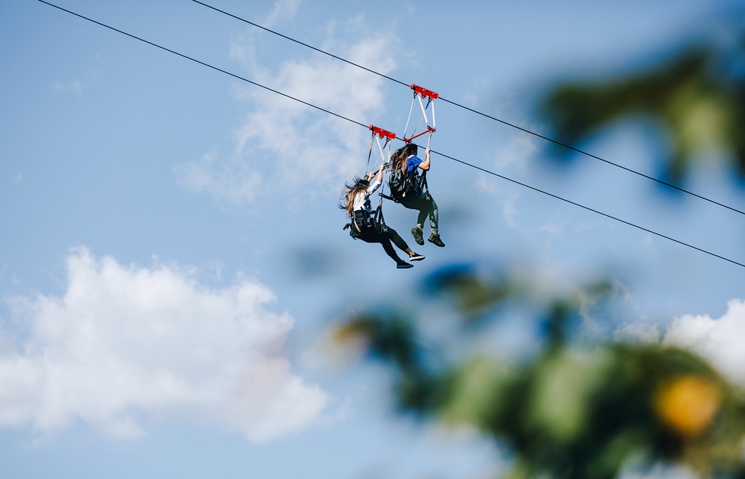 Two people ziplining against a blue sky with clouds.