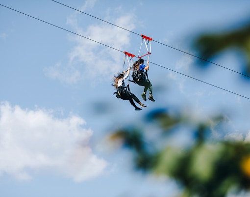 Two people ziplining against a blue sky with clouds.