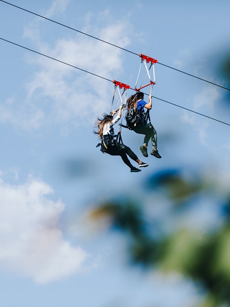 Two people ziplining against a blue sky with clouds.
