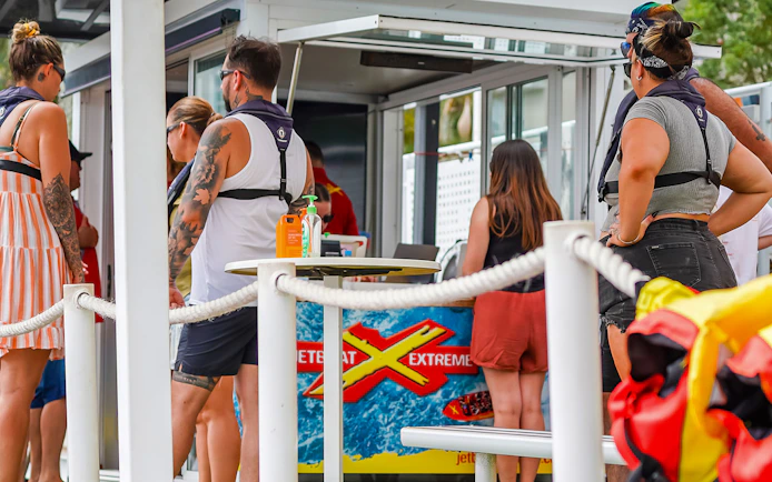 People preparing for the Ultimate Jet Boat Ride at a waterfront kiosk.