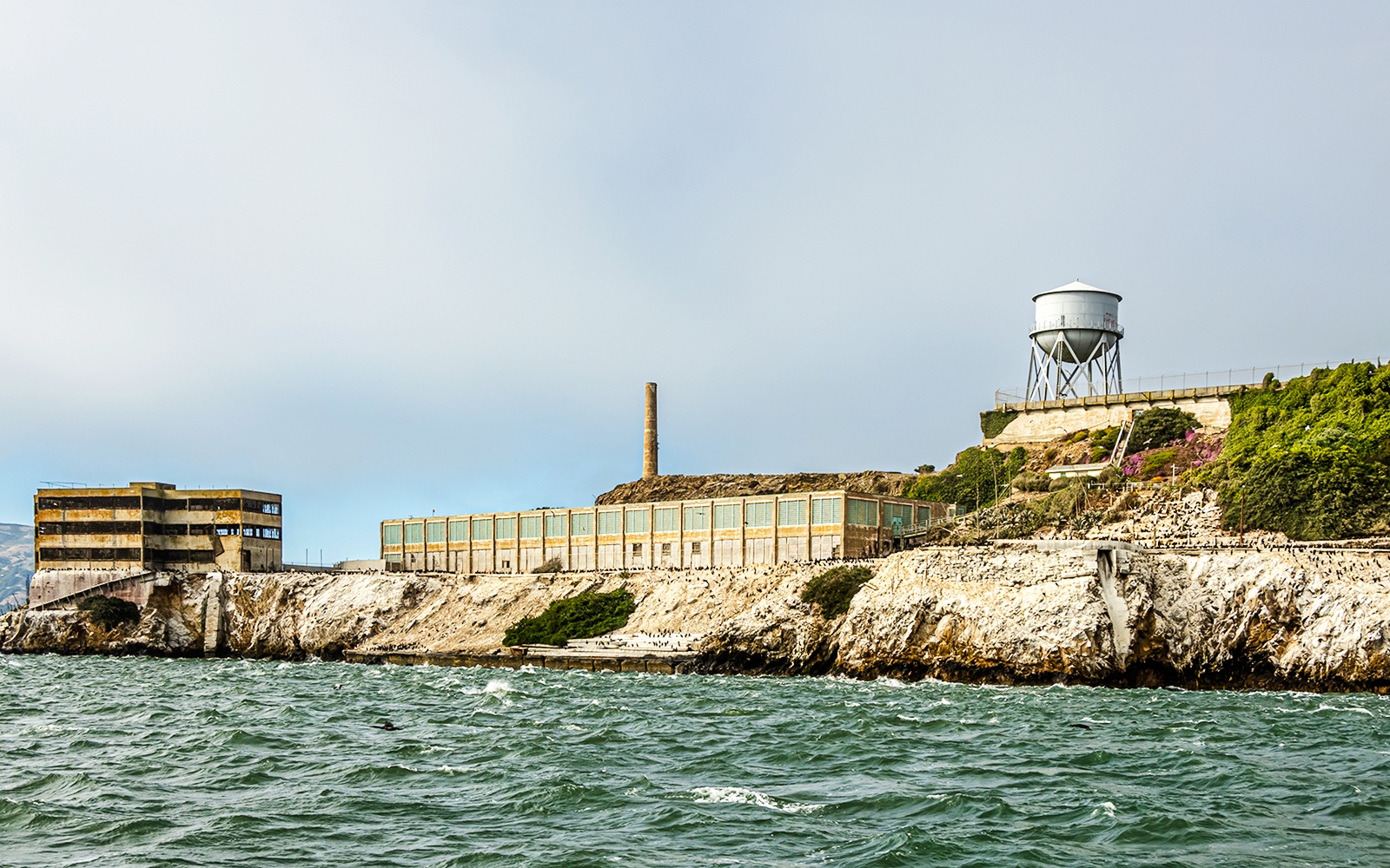 New Industries Building and water tower on Alcatraz Island, viewed from the water under a cloudy sky.