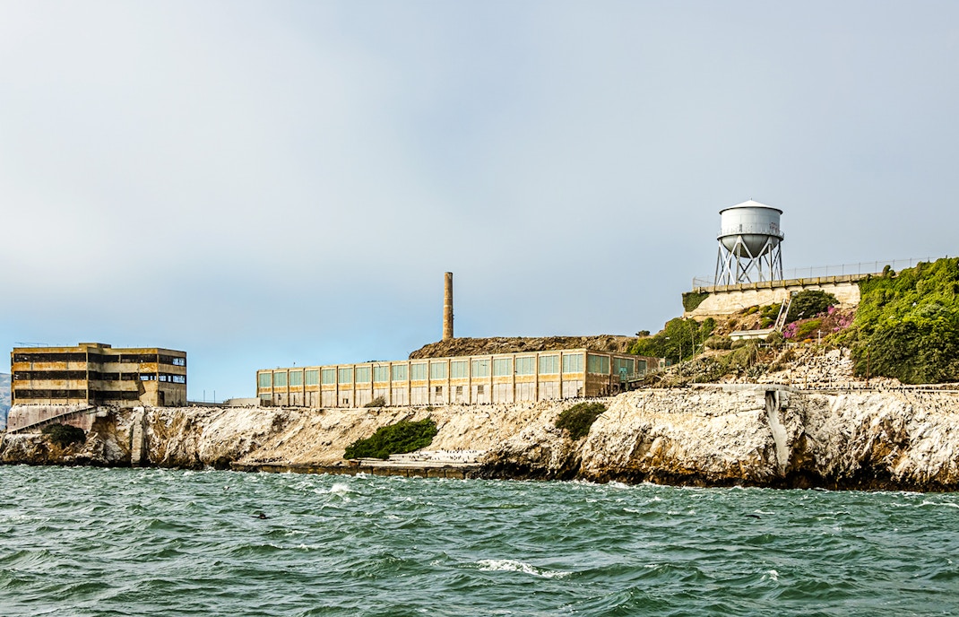 New Industries Building, Alcatraz Island from the water, showing the main prison buildings and a water tower on a rocky cliff under a cloudy sky