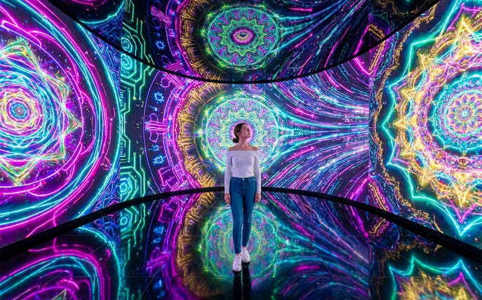 Woman standing amidst vibrant digital projections on walls and floor.