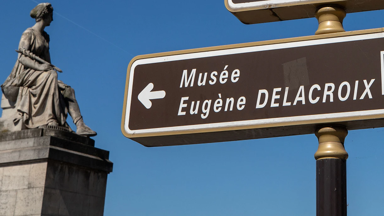 Sign pointing to Musée Eugène Delacroix with a statue in the background.