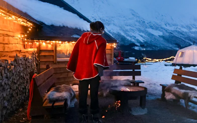 Person in red cloak by fire pit at snowy Tromso campsite with mountains and lights.