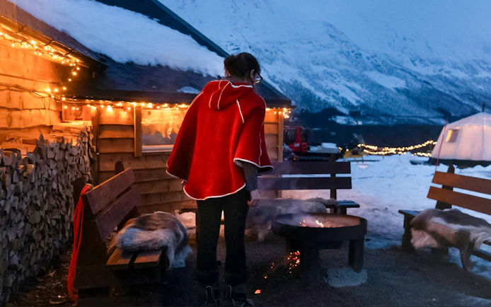 Person in red cloak by fire pit at snowy Tromso campsite with mountains and lights.