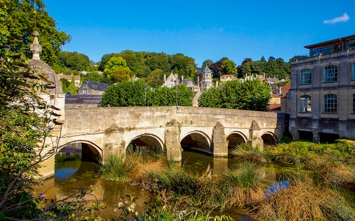 Historic bridge over river in Bath, England, surrounded by greenery and historic buildings.