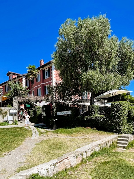 Lakeside view of a colorful building and church tower on Isole Borromee, Italy, with tourists walking nearby.