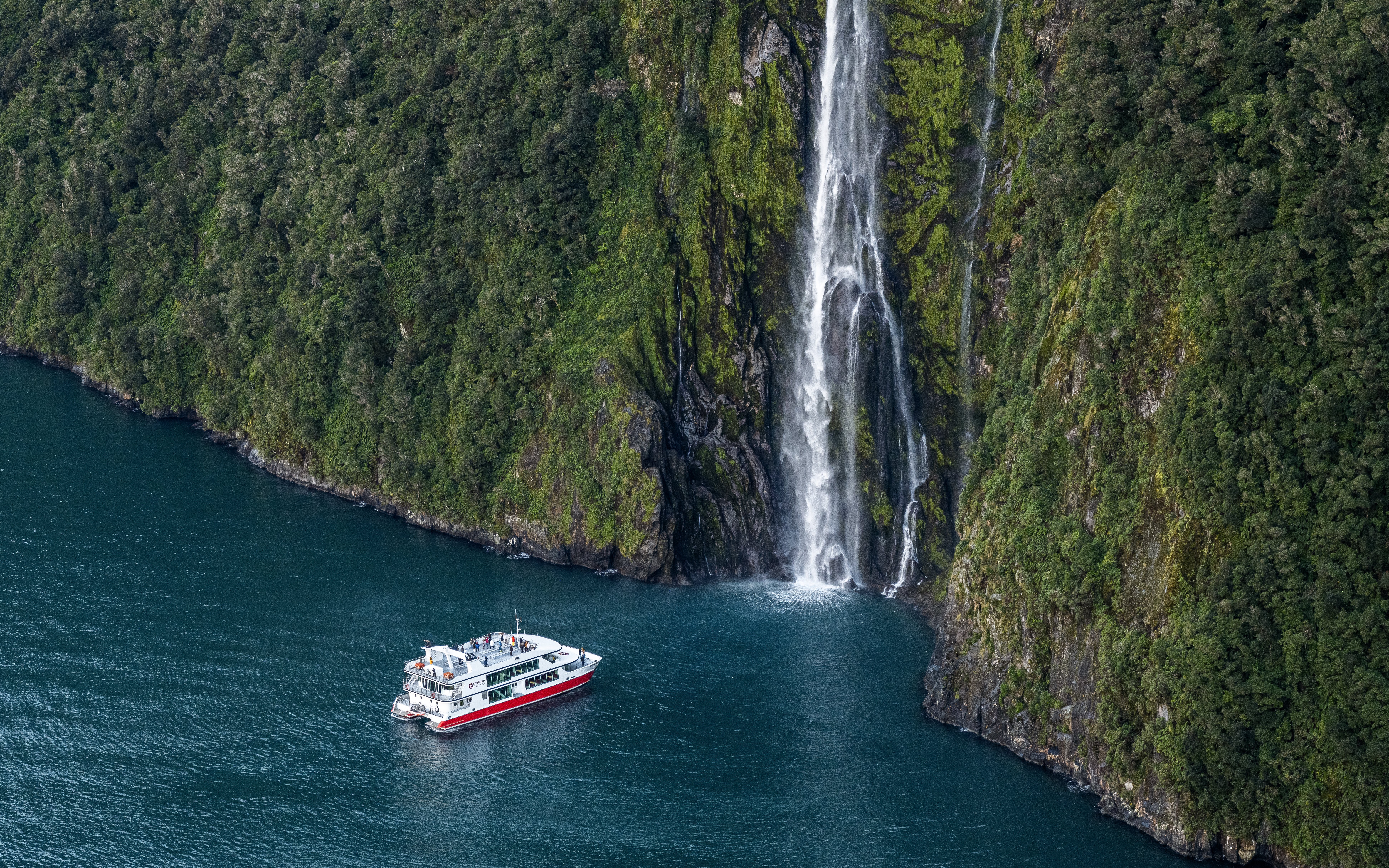 Cruise boat near waterfall in Milford Sound, New Zealand, surrounded by lush cliffs.