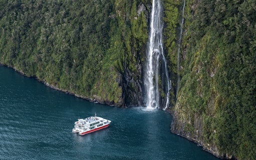 Cruise boat near waterfall in Milford Sound, New Zealand, surrounded by lush cliffs.