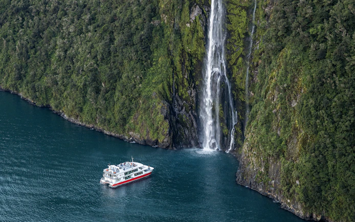 Cruise boat near waterfall in Milford Sound, New Zealand, surrounded by lush cliffs.
