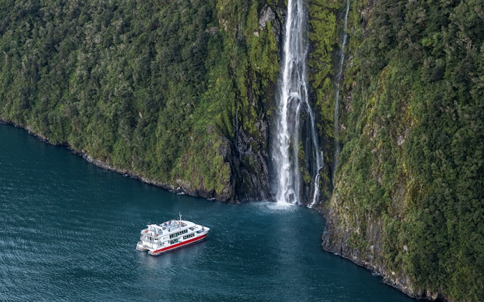 Cruise boat near waterfall in Milford Sound, New Zealand, surrounded by lush cliffs.